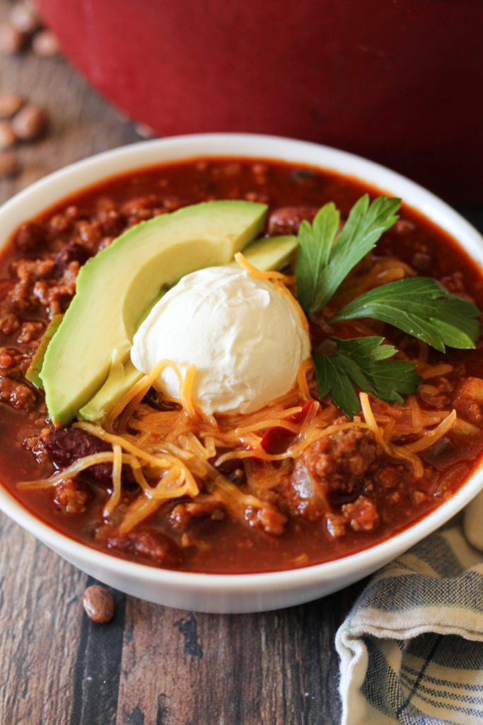 Homemade Chili is a family staple!! Loaded with veggies, ground beef and sausage and the perfect amount of heat. This easy chili recipe will warm you to the core. chili in a white bowl topped with avocado and sour cream