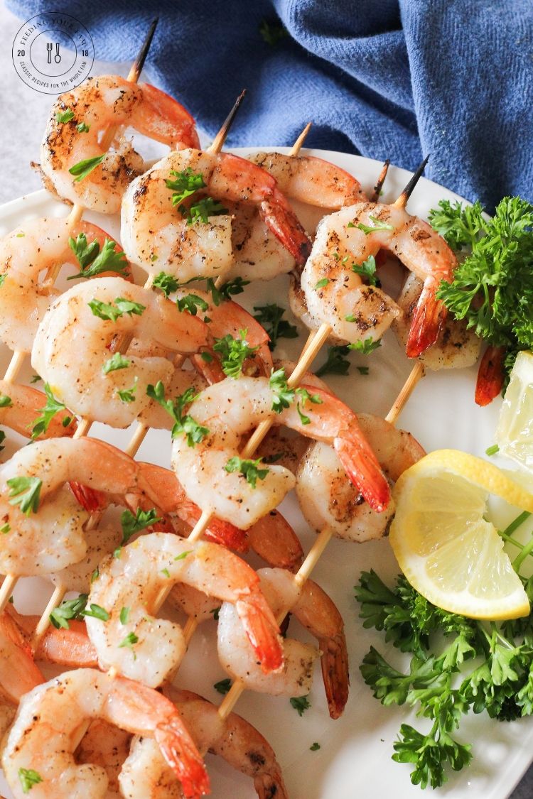 This easy Grilled Shrimp Recipe is a quick and simple meal that can be made on the grill with or without skewers. The ingredients are simple, complimentary and minimal. These grilled shrimp will be a repeat on your menu! #grilledshrimp #grillingrecipes #shrimprecipes grilled shrimp skewers on a white plate with lemon and parsley