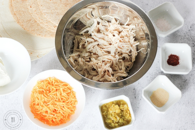 Ingredients for chicken taquitos including shredded chicken, cream cheese, green chiles, shredded cheddar, and spices on a countertop