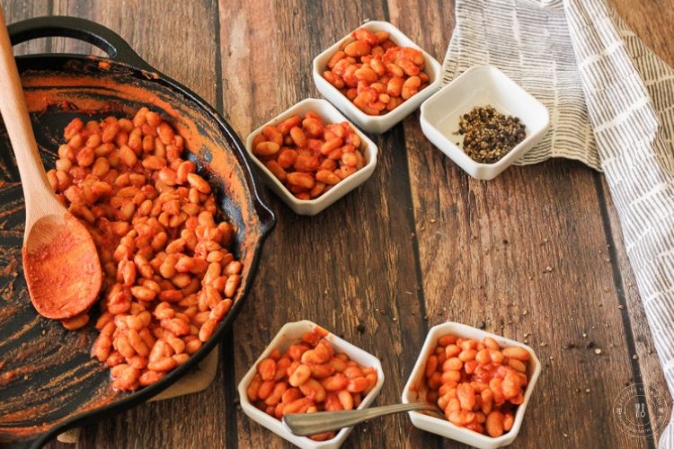 I know you've heard of Buffalo Chicken Wings, now prepare your palette for these zippy BUFFALO BAKED BEANS! Buffalo Baked Beans are a perfect combination of tangy and heat. These beans pack a tastebud punch!! #bakedbeans #buffalobakedbeans #bbqsidedish buffalo baked beans in a cast iron skillet next to bowls of beans