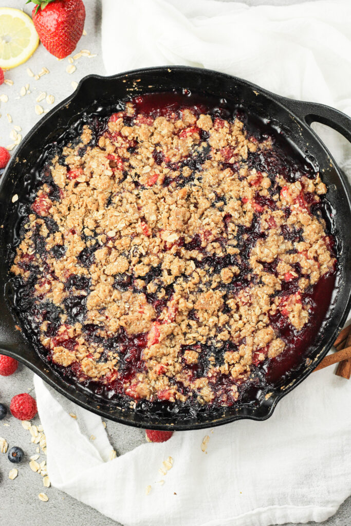 triple berry crisp with cooked berries topped with an oatmeal crips topping in cast iron pan