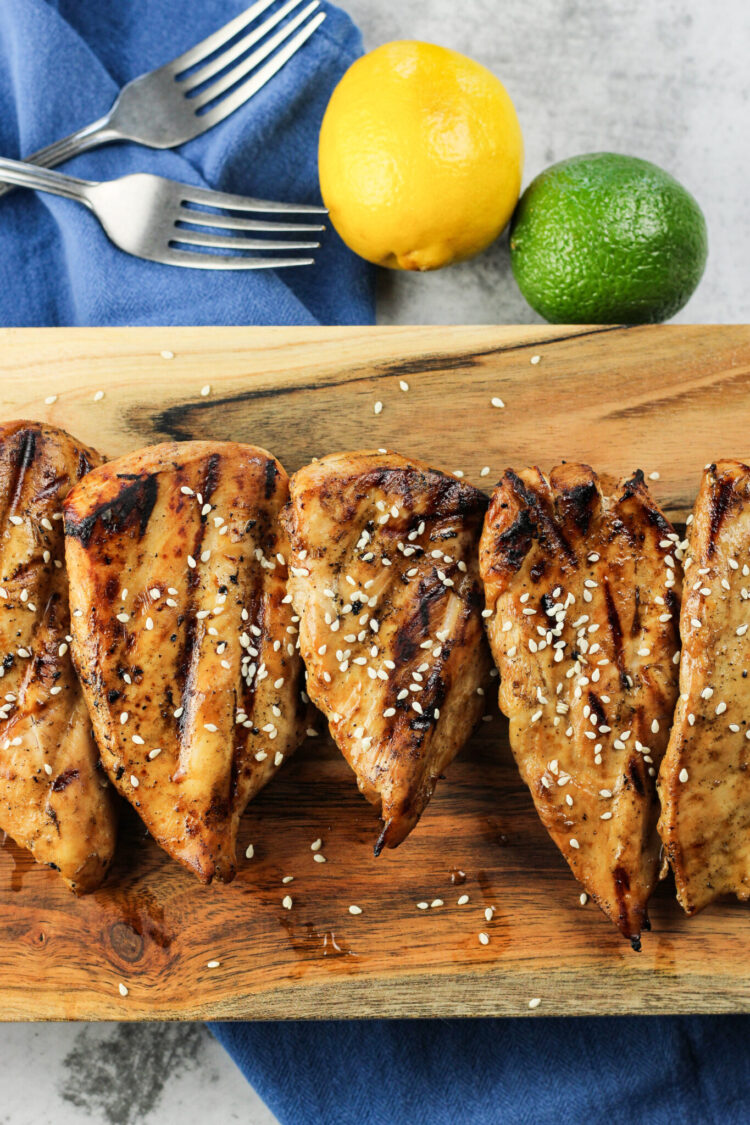 grilled teriyaki chicken breasts on a wooden board with forks and a lemon and lime on the side