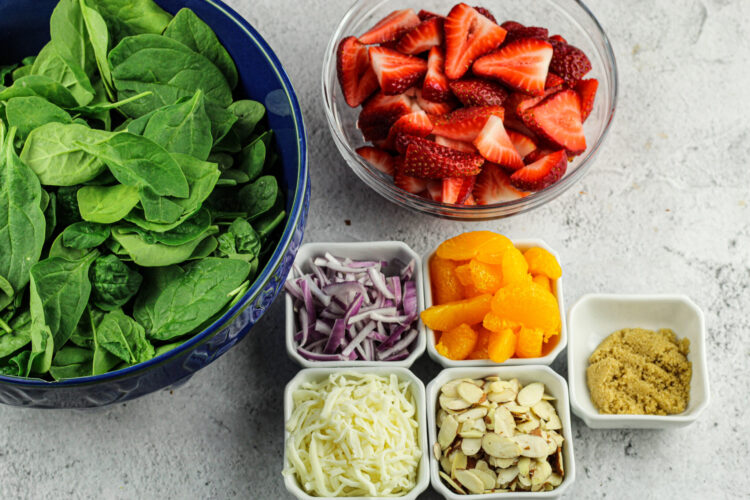 big bowl of spinach, bowl of sliced strawberries, mandarin oranges, sliced red onions, mozzarella cheese, almonds and brown sugar