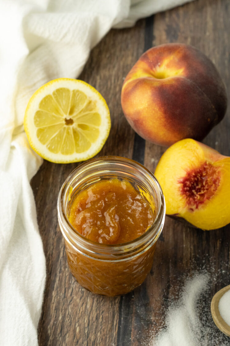 peach jam in a glass jar surrounded by peaches and a sliced lemon