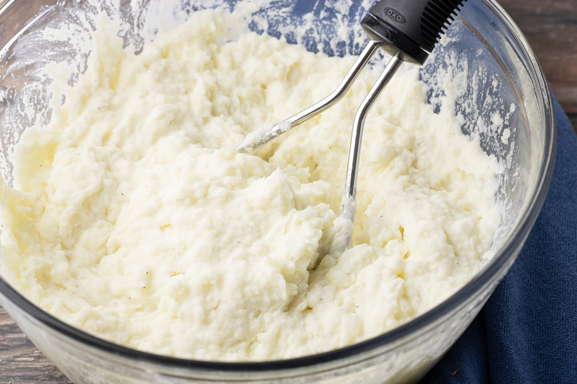 mashed potatoes in a mixing bowl with a potato masher