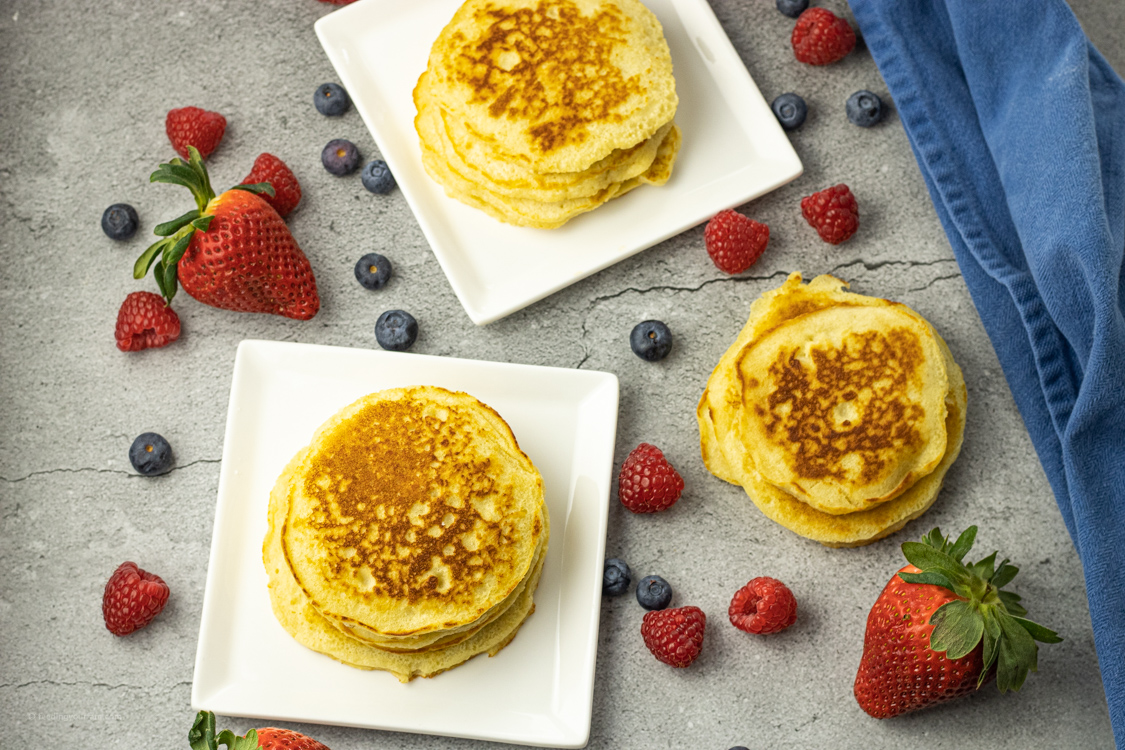 piles of cooked pancakes on white plates surrounded by berries