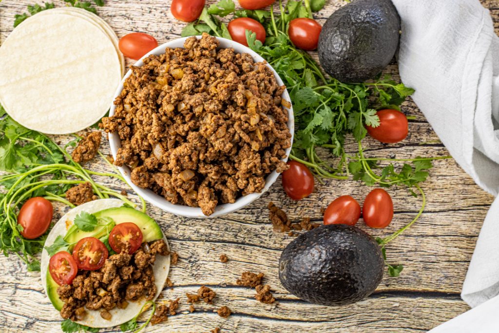 bowl of ground beef taco meat surrounded by avocados, cilantro and grape tomatoes