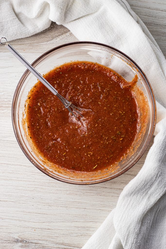 glass bowl of tomato sauce mixed with herbs and spices with a mini whisk