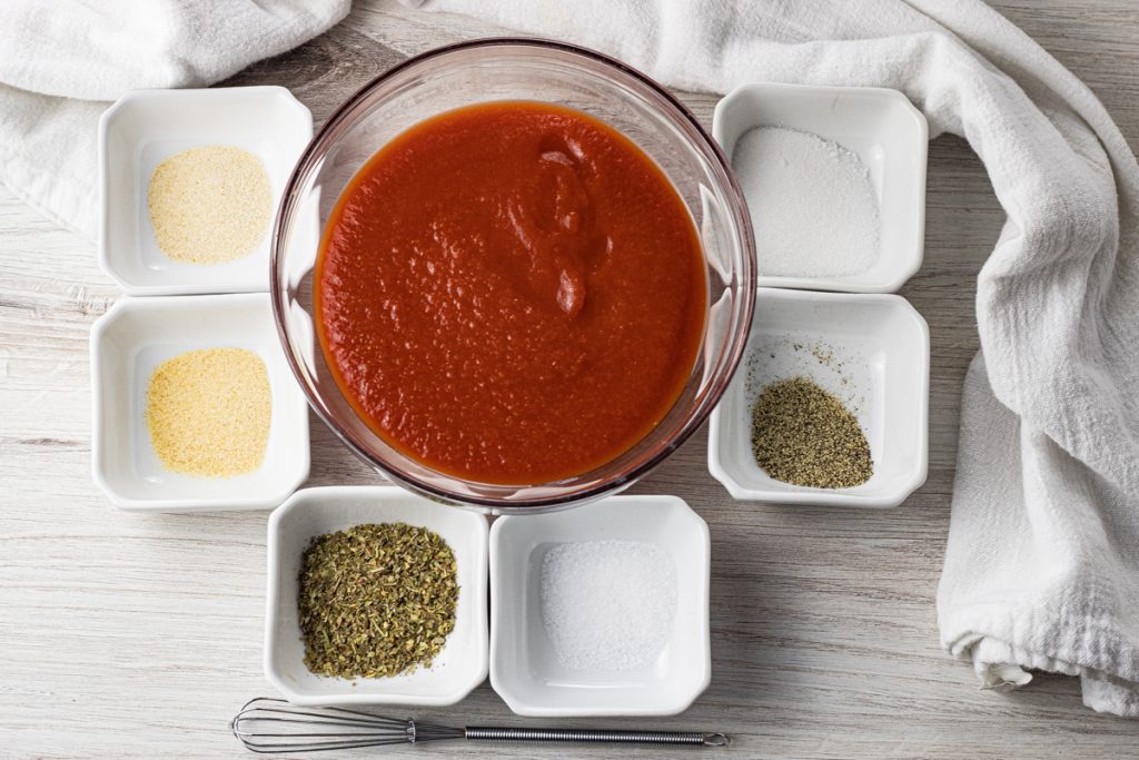 big glass bowl of tomato sauce surrounded by smaller white bowls with herbs and spices in them