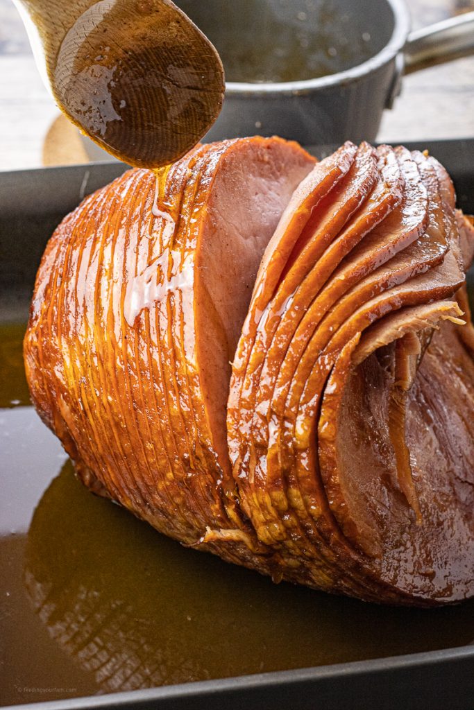 sliced ham being drizzled with brown sugar glaze in a roasting pan