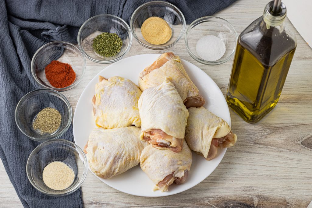 chicken thighs with skin on surrounded by small bowls filled with spices and a jar of olive oil