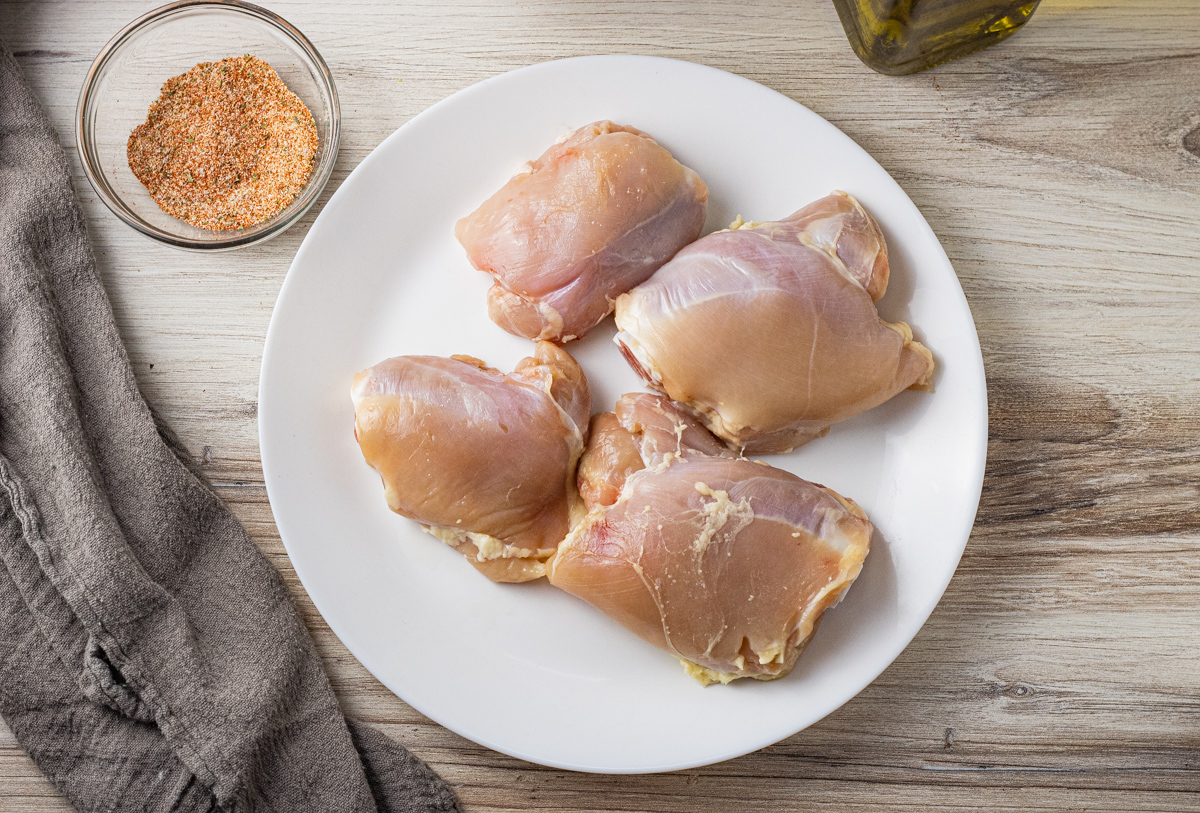 skinless, bone in chicken thighs on a white plate with a bowl of spices next to it