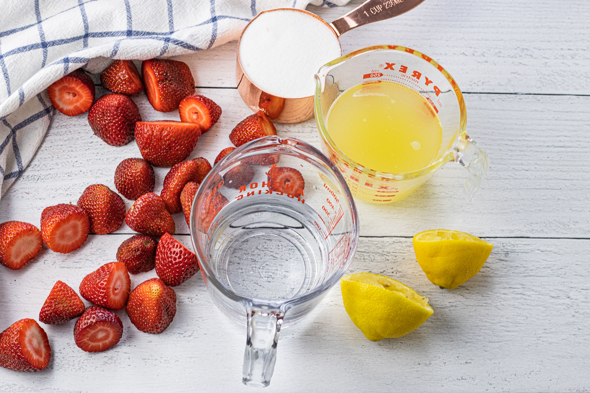 sliced strawberries, a cup of sugar, water and lemon juice with lemons that have been squeezed. 