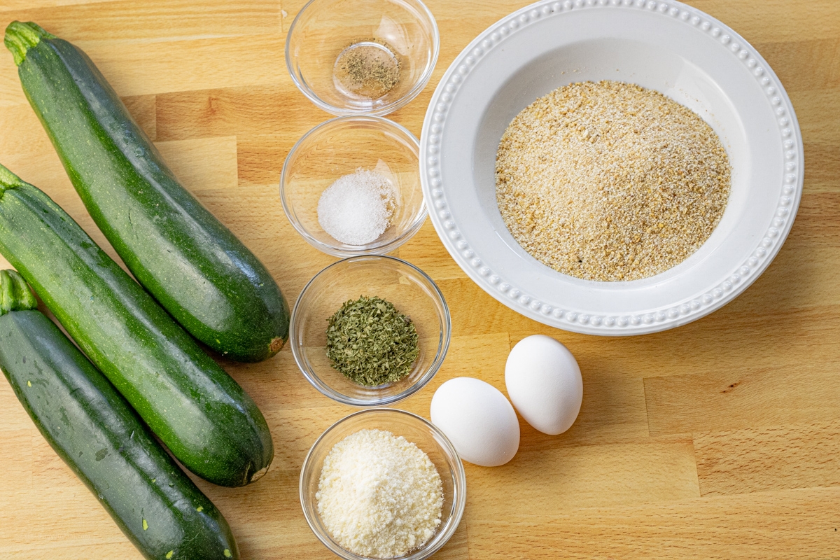 bowl of bread crumbs, two eggs, small bowls of spices and parmesan cheese and three green zucchini