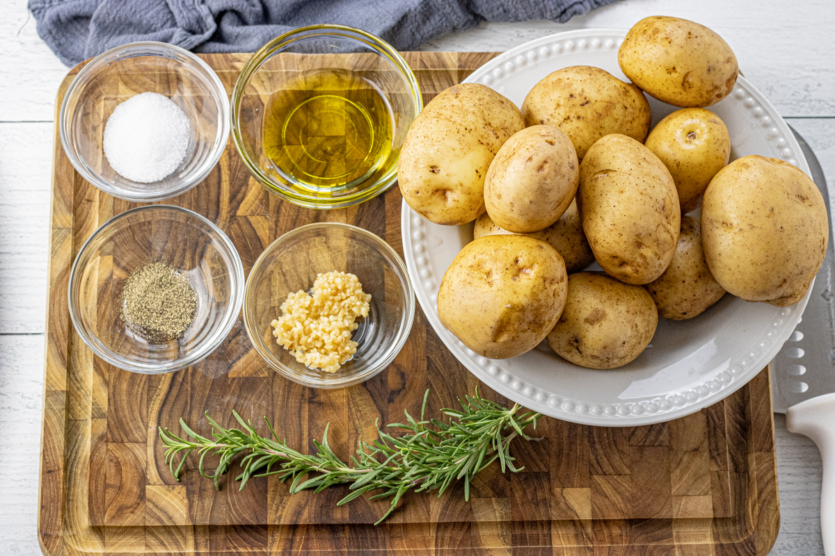 bowls of garlic, salt, pepper, olive oil and golden potatoes and a sprig of rosemary