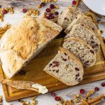 Sliced loaf of cranberry walnut bread on a wooden cutting board with dried cranberries and walnuts.