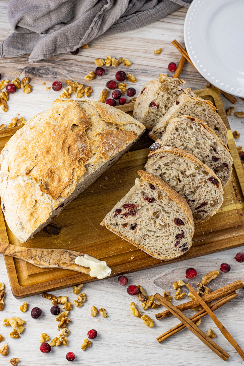Sliced loaf of cranberry walnut bread on a wooden cutting board with dried cranberries and walnuts.