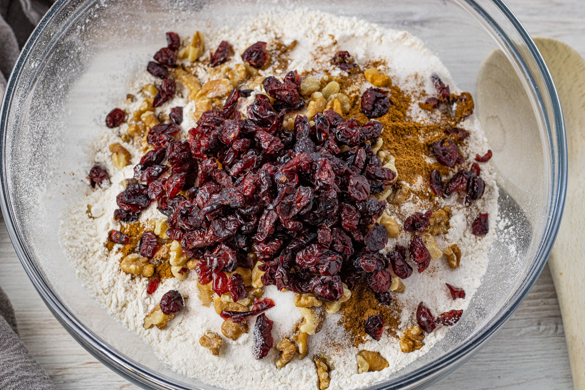 Dry bread ingredients including flour, cinnamon, dried cranberries, and chopped walnuts mixed in a glass bowl.