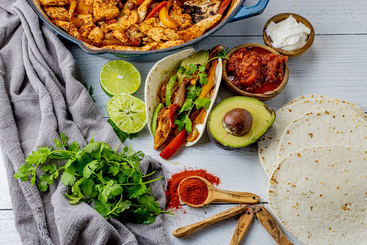 chicken fajita surrounded by limes, avocados, flour tortillas, salsa, sour cream and cilantro