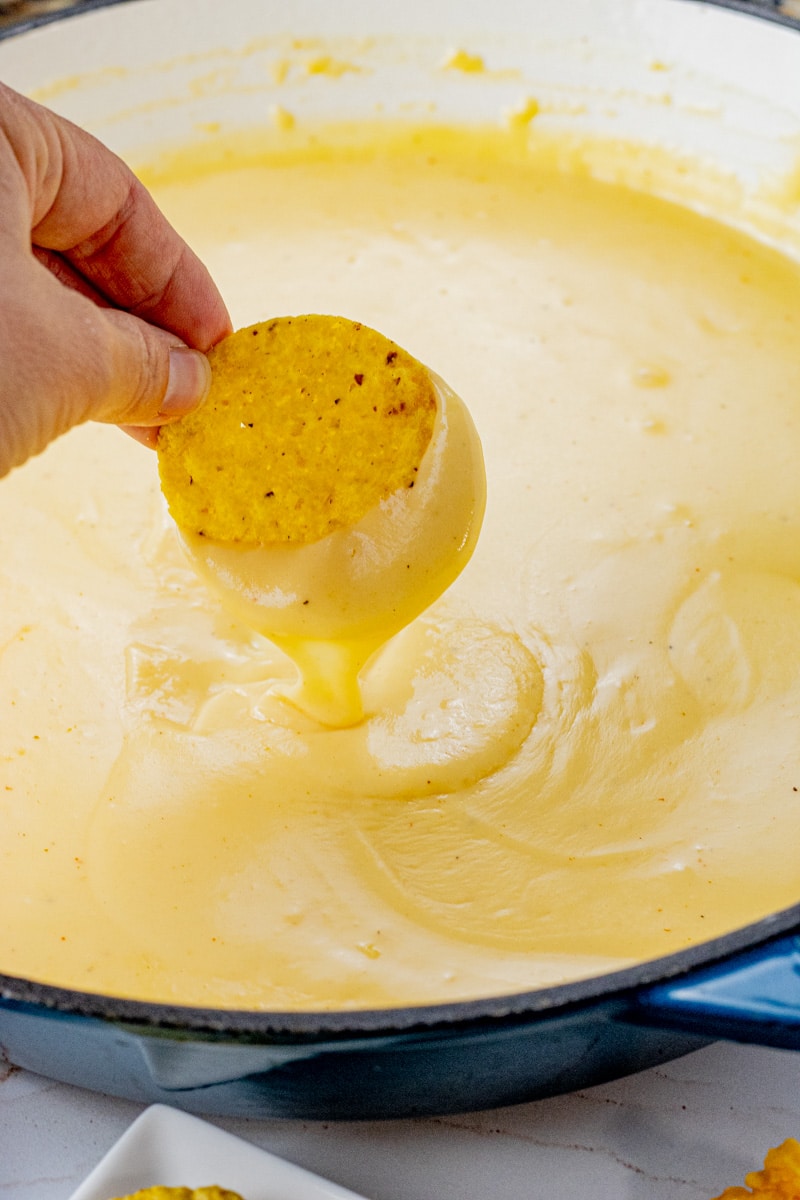 hand holding a round tortilla chip with cheese sauce dripping from the chip into a pan of cheese sauce