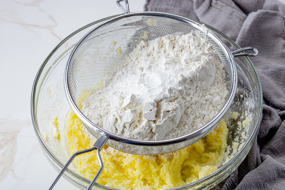 flour in a mesh strainer over creamed butter and sugar in a glass mixing bowl