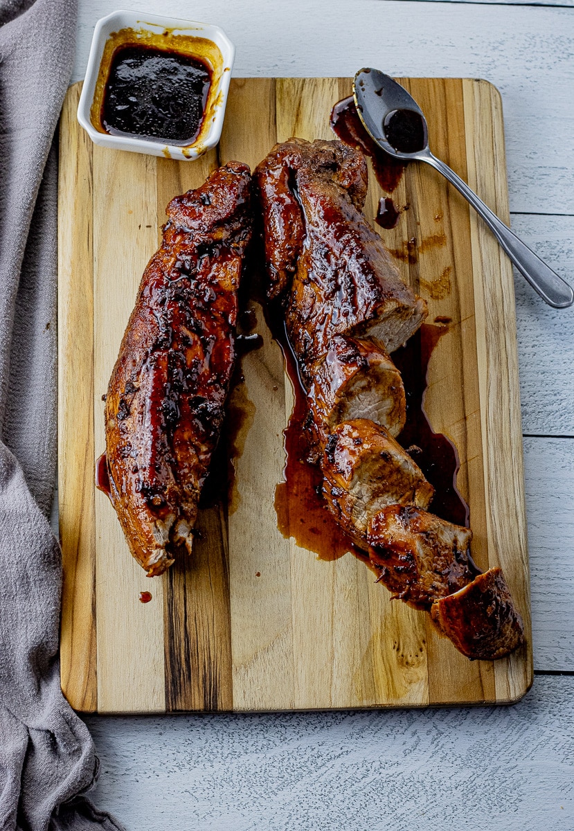 two pork tenderloins on a wooden cutting board, one sliced and drizzled with a maple glaze