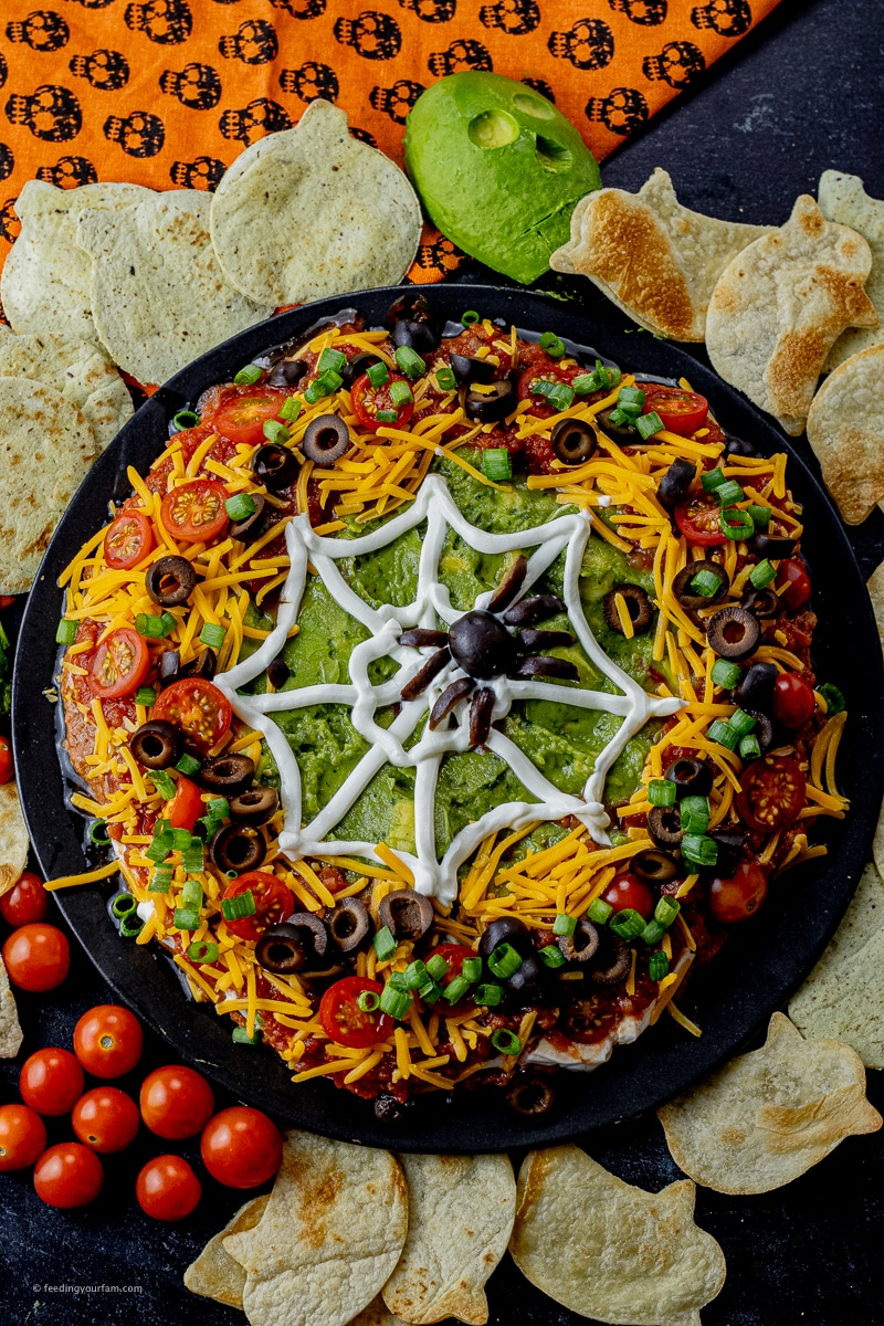 taco dip made with layers of beans, cheese, salsa, guacamole, olives and tomatoes, topped with a sour cream spider web and a spider made out of an olive.