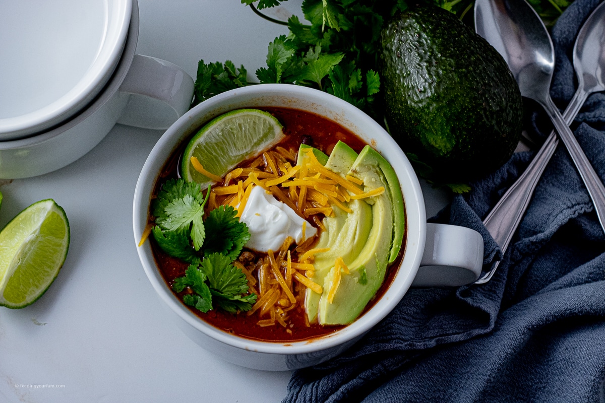 bowl of taco soup topped with cilantro, cheese, lime and sliced avocado