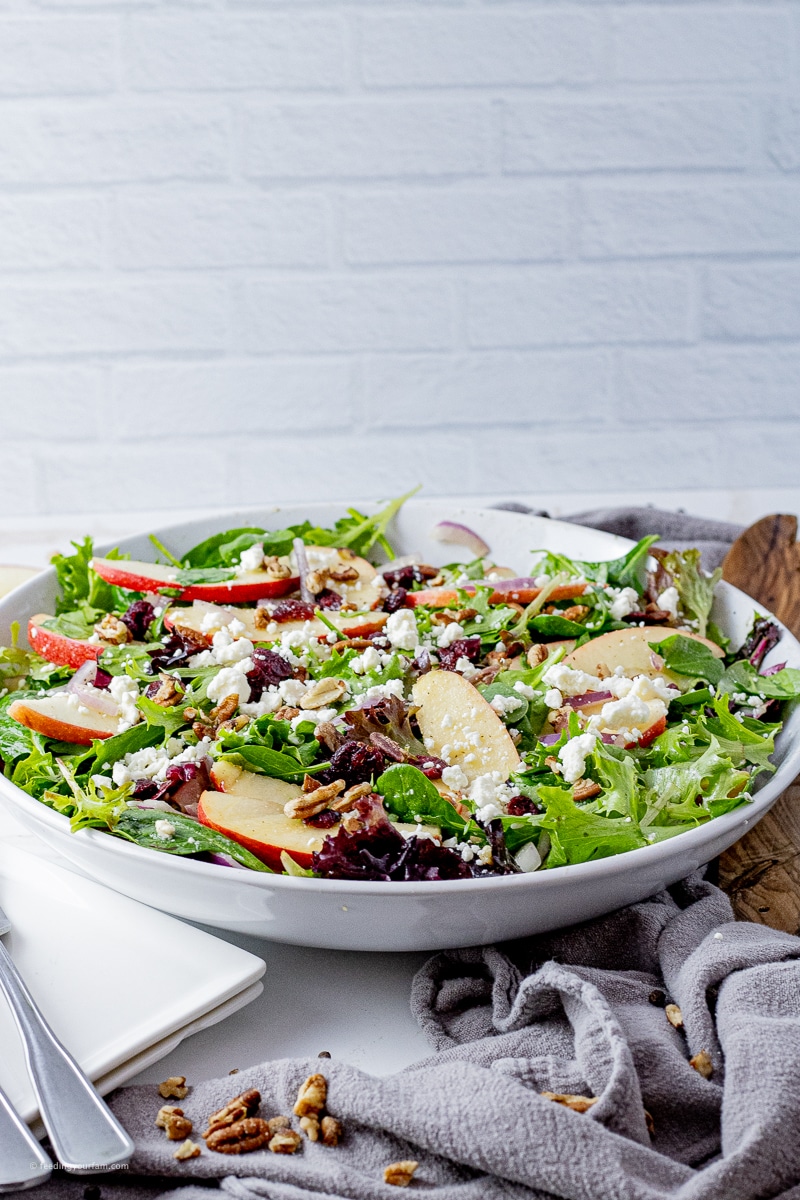 big white bowl filled with a spinach salad with sliced apples, red onion slices, pecans and feta cheese