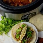 pulled pork in a slow cooker with some tacos outside on a white plate