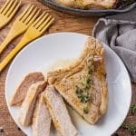 bone in pork chops cooking in a cast iron pan, one is on a white plate and sliced into smaller slices.
