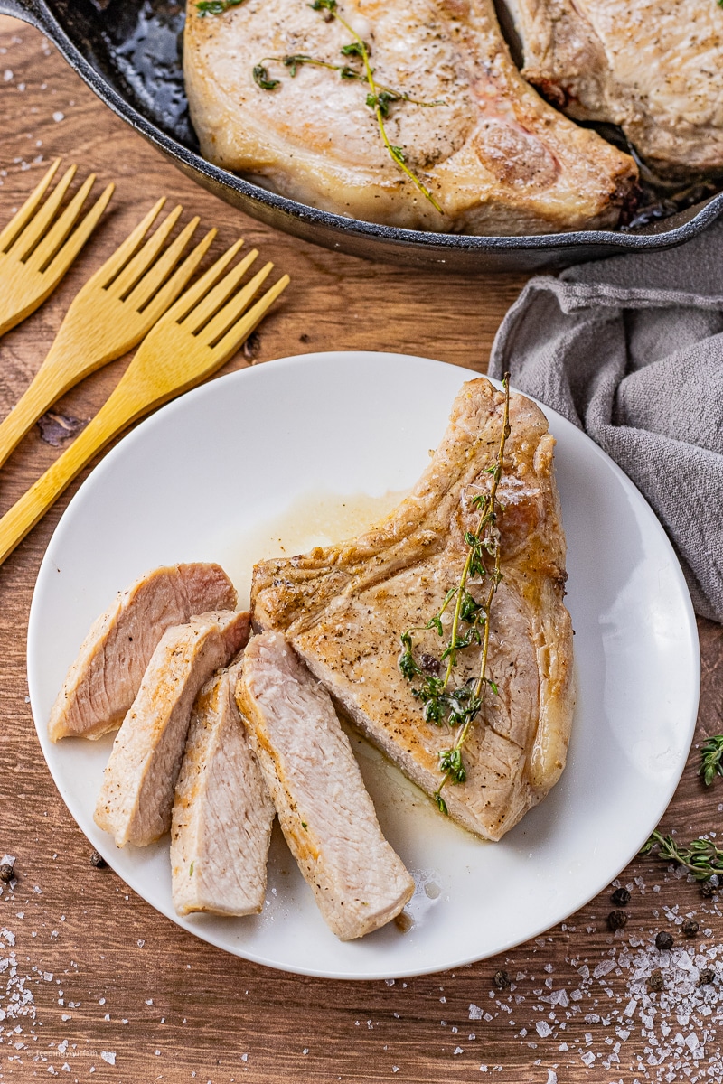 bone in pork chops cooking in a cast iron pan, one is on a white plate and sliced into smaller slices.