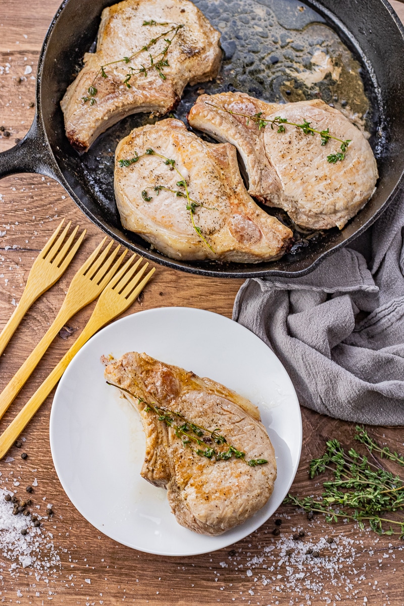 bone in pork chops cooked in a cast iron pan, one is on a white plate with a sprig of thyme on top