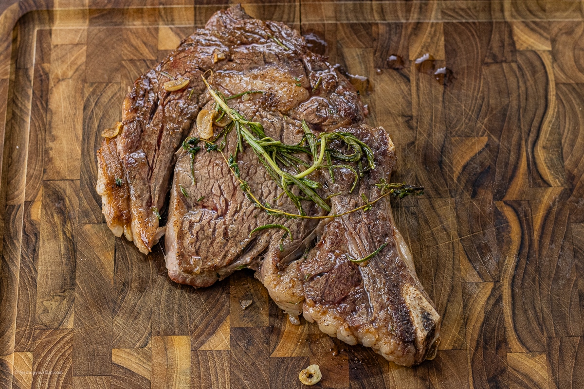 cooked ribeye steak on a wooden cutting board with a sprig of rosemary on top of the steak