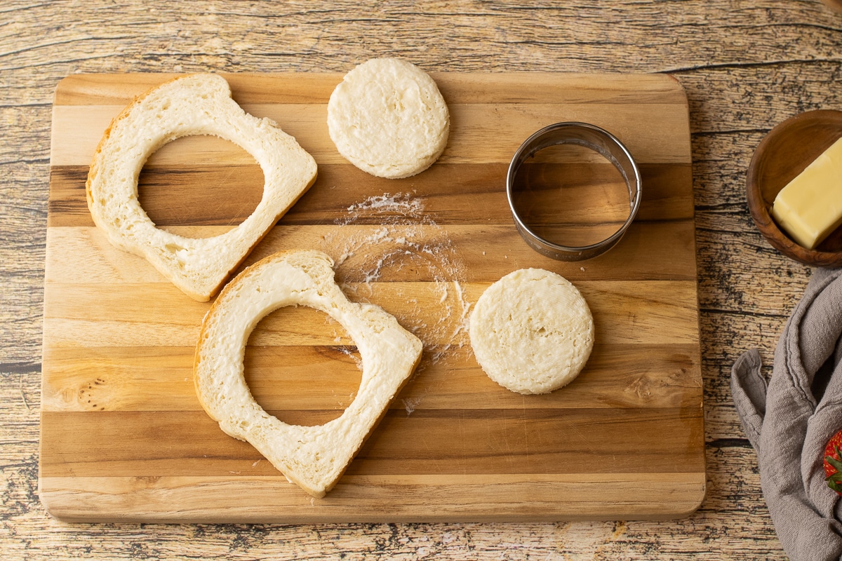 white bread buttered with circles cut out of the center on a wooden cutting board