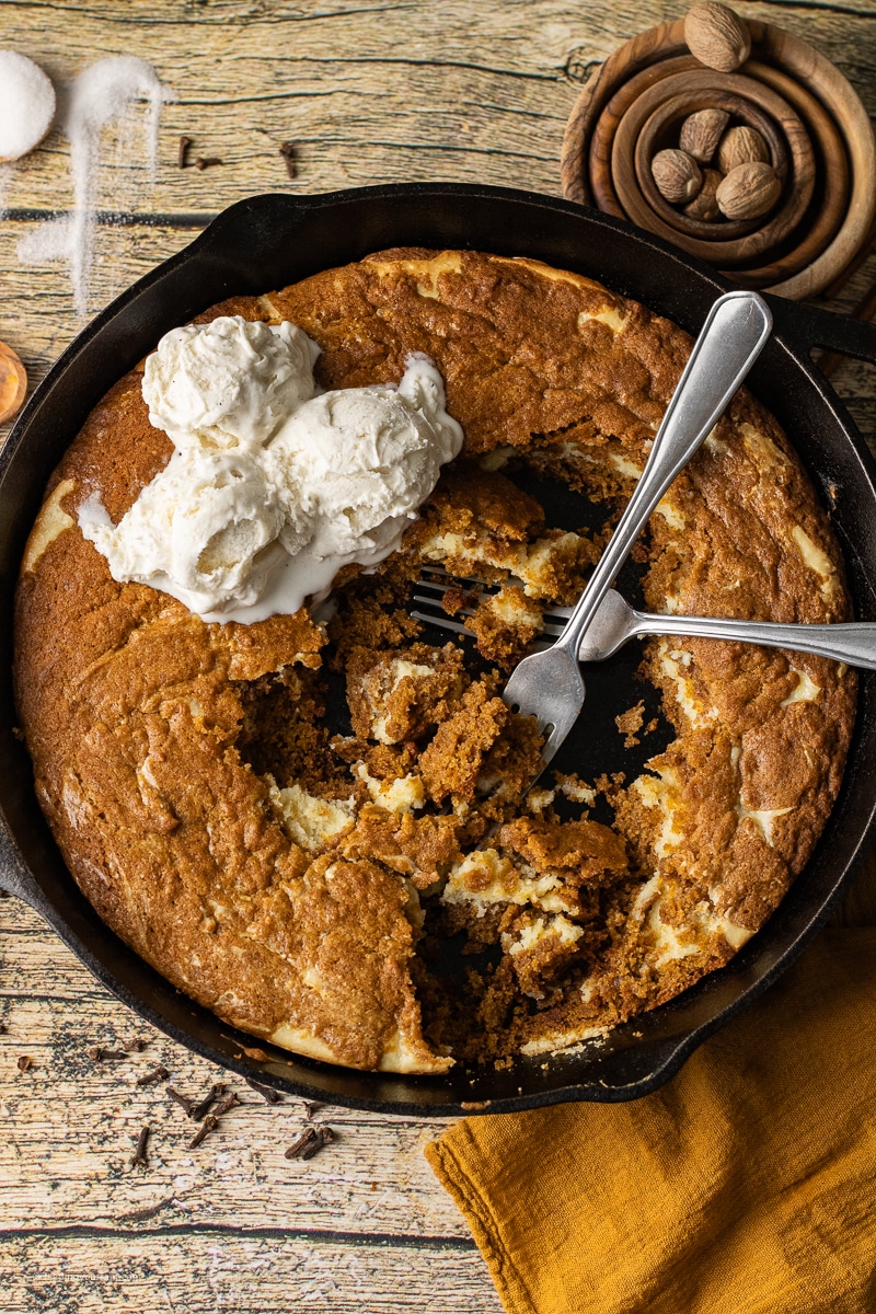 A cast iron skillet filled with a golden pumpkin cookie swirled with creamy cheesecake, baked until soft in the center with crisp edges