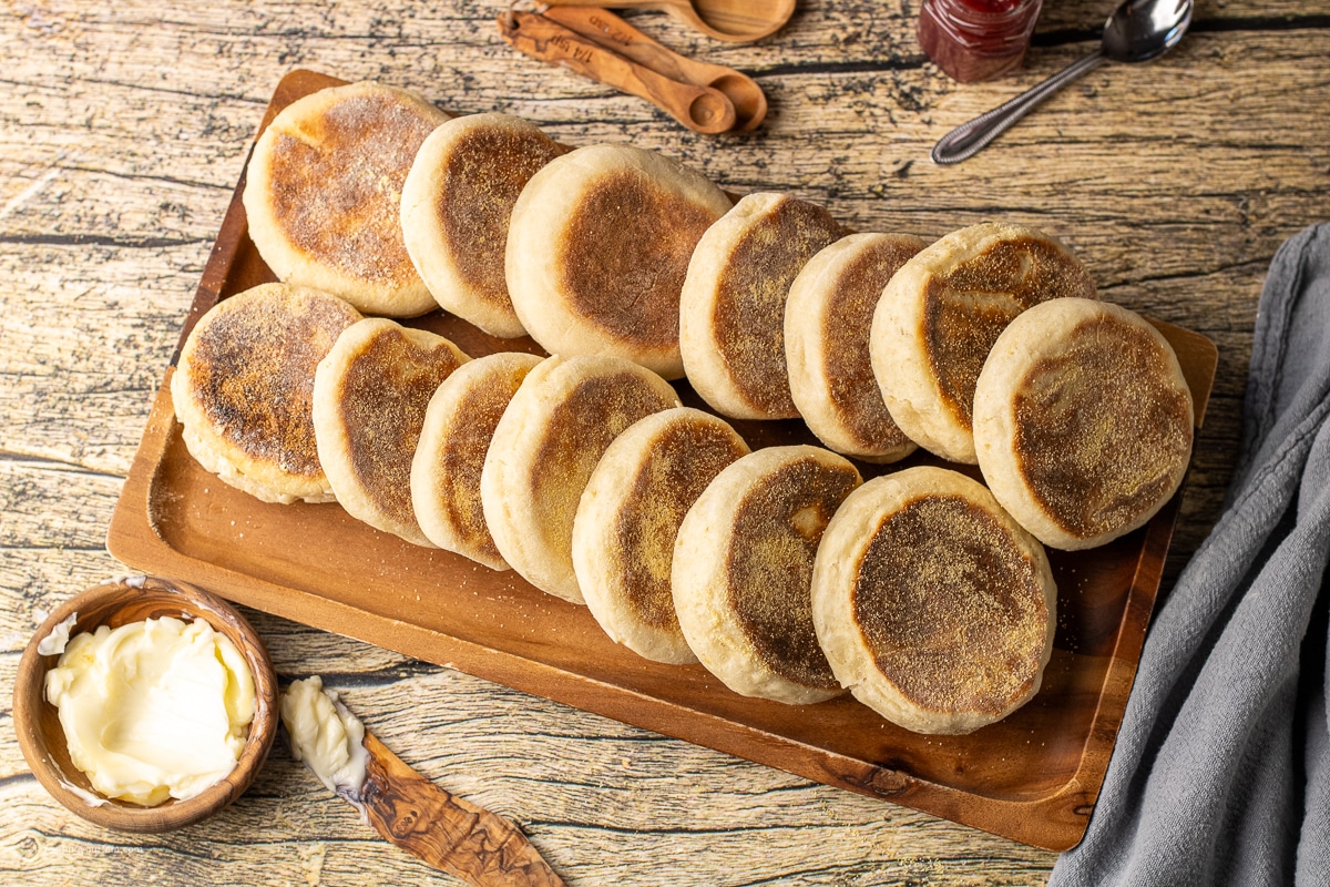 homemade english muffins cooked on the stove top in a cast iron skillet until golden brown then placed on a wooden serving platter. 